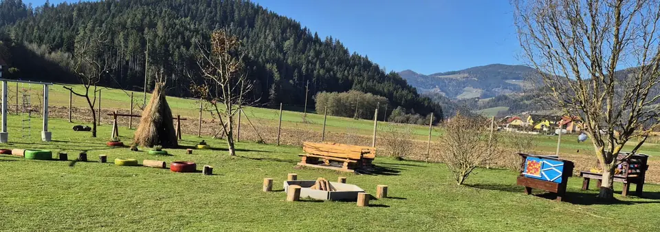 Travnik igralnice v naravi Lalalu s klopmi, ognjiščem in leseno hiško, obdan z gozdom